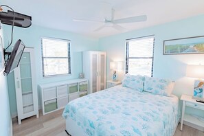 A bedroom with a bed, two windows with blinds, a ceiling fan, a desk with drawers, and a nightstand with a lamp. - Serenity by the Sea, Beach Condo, w/Pool & Access (St. Pete Beach)
