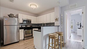 A modern kitchen with white cabinets, stainless steel appliances, a central island with stools, and a view into adjacent rooms. - Lovely Water View Condo | Top Floor | Steps to Beach (Madeira Beach)