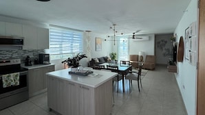 Ett modernt kök med en köksö i mitten, en matplats med bord och stolar samt ett vardagsrum med soffa och fåtöljer.