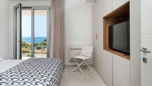 A modern bedroom with a bed, a TV mounted on a wooden panel, and a sliding glass door leading to a balcony with a view of the sea. - Villa Silvia with swimming pool - My Rental Homes (Syracuse)
