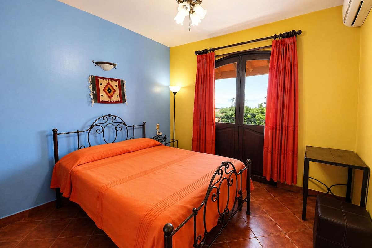 Un dormitorio con cama, mesita de noche, lámpara, ventana con cortinas y aire acondicionado.