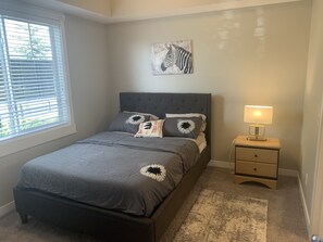 A bedroom with a bed, a bedside table with a lamp, a window with blinds, and a zebra painting on the wall.