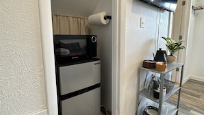 Eine kleine Küche mit Kühlschrank, Mikrowelle und einer kompakten Arbeitsfläche. Ein Flachbildfernseher ist über der Arbeitsfläche angebracht.