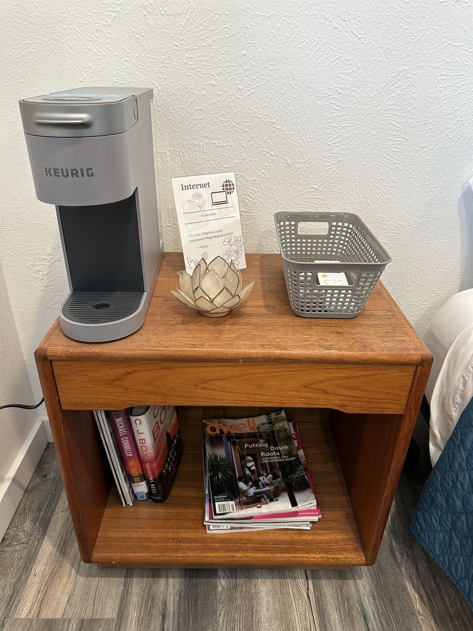 A wooden nightstand with a Keurig coffee maker, a basket, a card, and magazines.