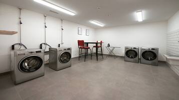 A laundry facility with four washing machines, a table, two chairs, and a wall-mounted ironing board.