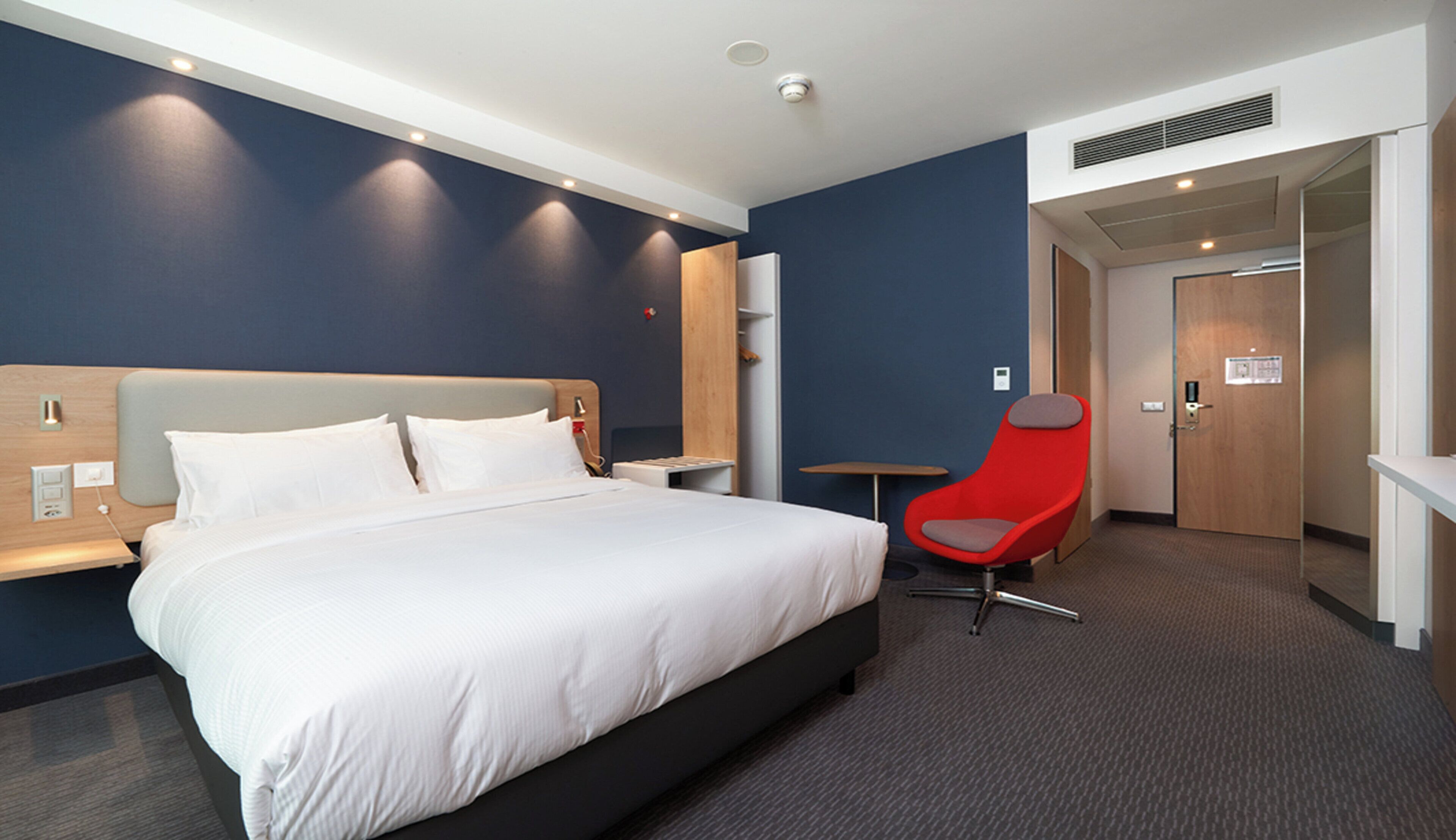 Ett hotellrum med en stor säng, en röd stol, ett litet bord och en dörr som leder till ett annat rum.