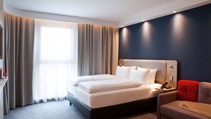 Habitación de hotel moderna con una cama grande, una mesita de noche con lámpara, una silla roja y una pequeña mesa de madera.
