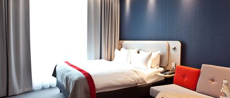 A modern hotel room with a large bed, a sofa, a small table, and a view of the outdoors.