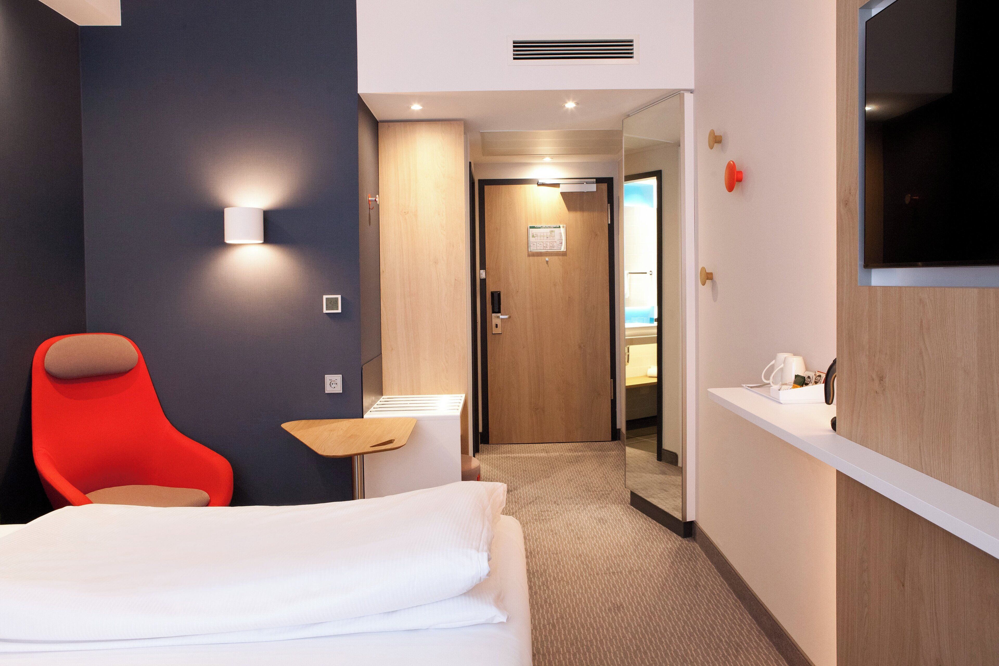 A hotel room with a bed, a red chair, a small table, a TV, and a door leading to a bathroom.