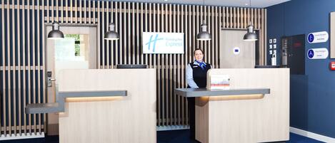 A Holiday Inn Express reception area with two reception desks, a blue wall with vertical wooden slats, and a sign indicating the General Manager's office.