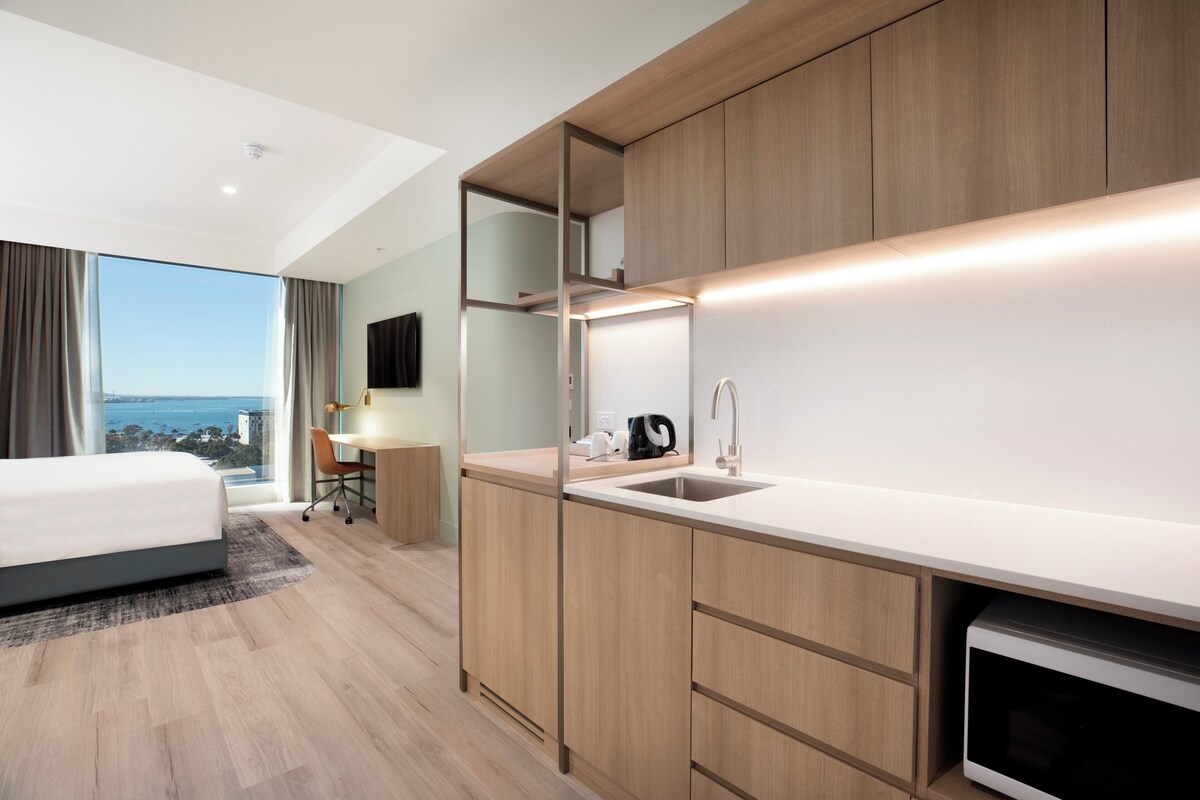 A modern hotel room with a large bed, a desk, and a kitchenette.