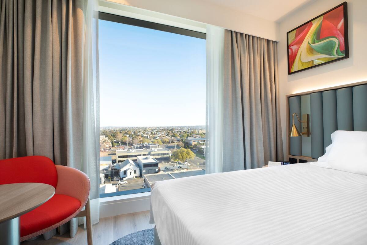 A hotel room with a large bed, a red chair, a table, and a city view through the window.