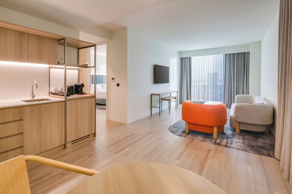 A modern hotel room with a wooden kitchenette, a seating area with an orange ottoman, a flat-screen TV, and a large window with curtains.