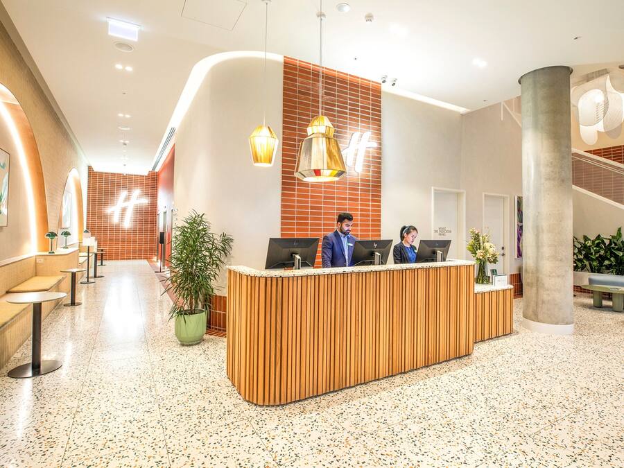 Una zona de recepción moderna de hotel con un mostrador de madera, dos recepcionistas, una planta grande en maceta y una escalera con barandilla.