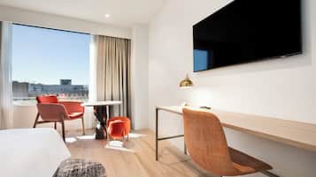A hotel room with a bed, a desk, a chair, a TV, and a window with a city view.