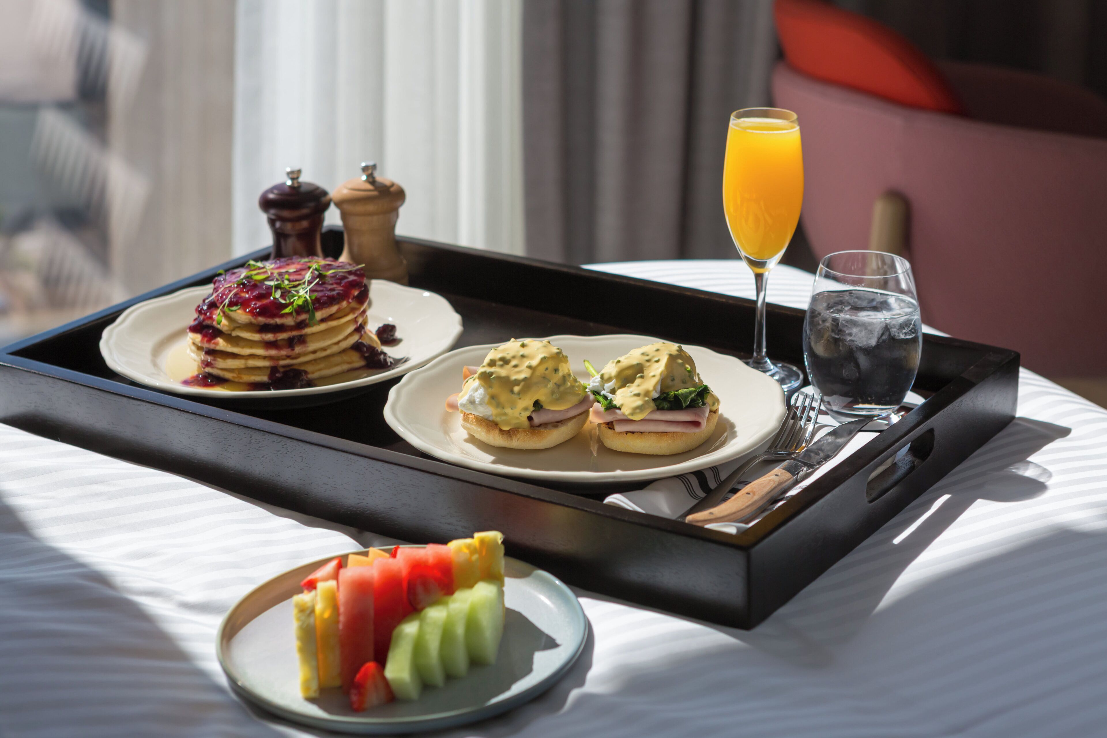 A breakfast tray with pancakes, eggs, and juice on a hotel bed.