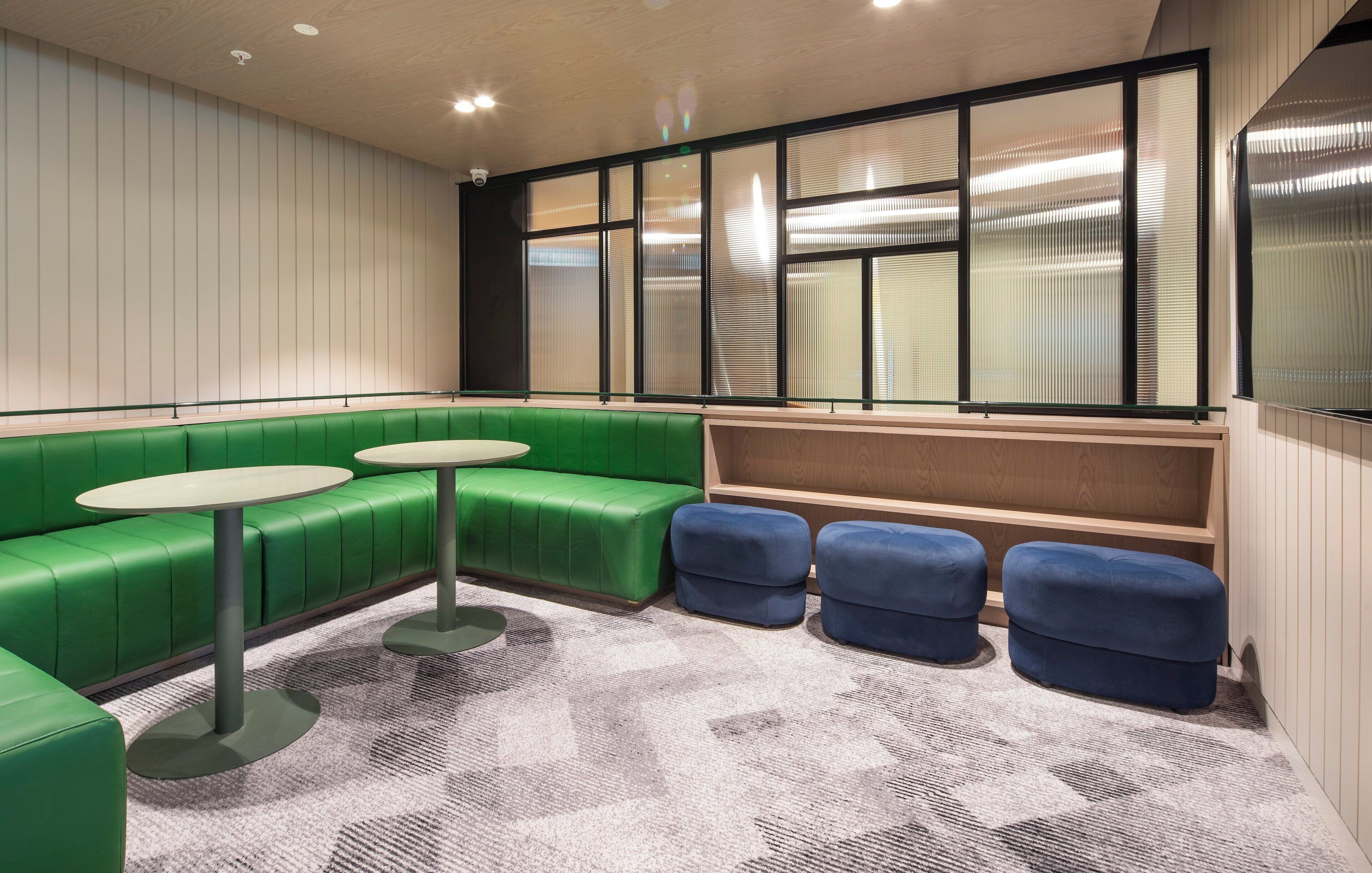 A modern lounge area with green seating, a round table, and a large window with blinds.
