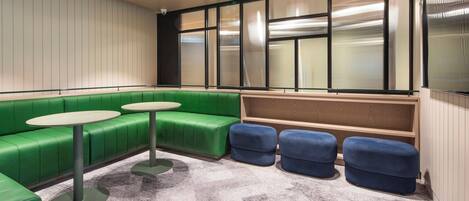 A modern lounge area with green seating, a round table, and a large window with blinds.