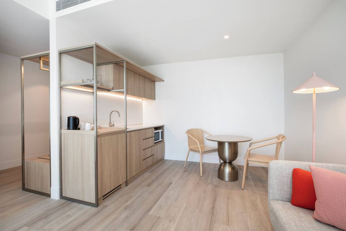 A modern kitchen with wooden cabinets, a microwave, and a kettle on the counter. There is a dining table with two chairs and a floor lamp.