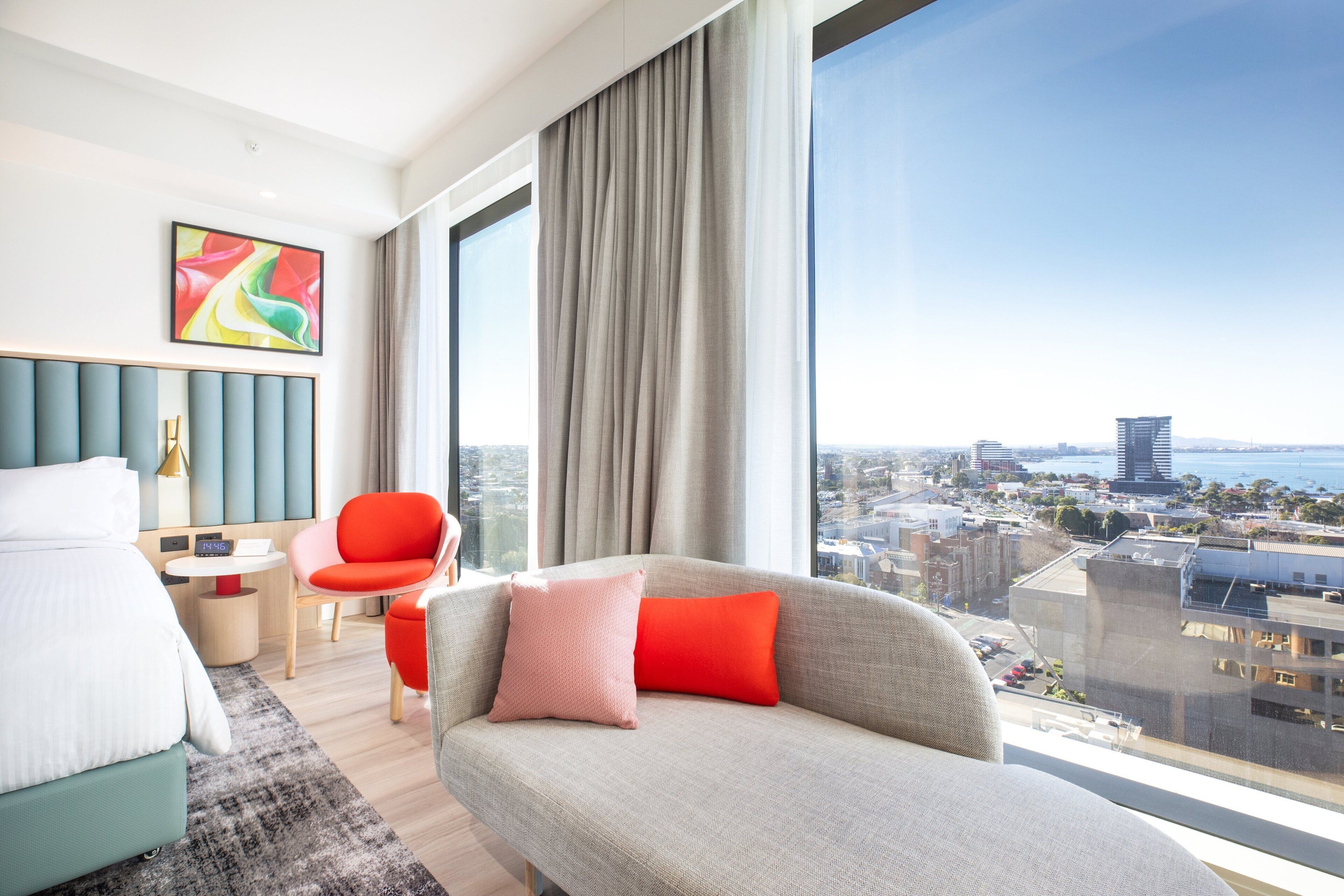 A modern hotel room with a large window offering a city view, a comfortable sofa, a dining area, and a bed.