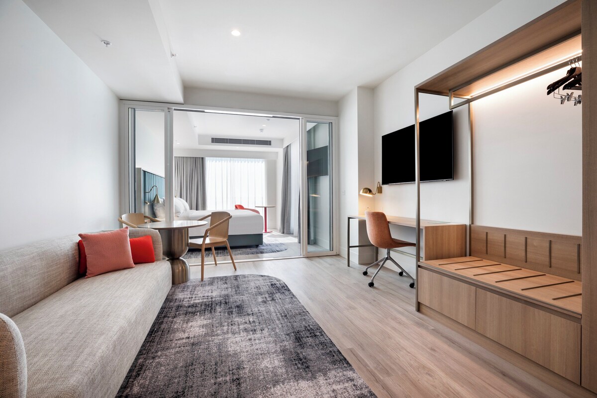A modern hotel room with a sofa, a desk, a chair, a TV, and a wardrobe.