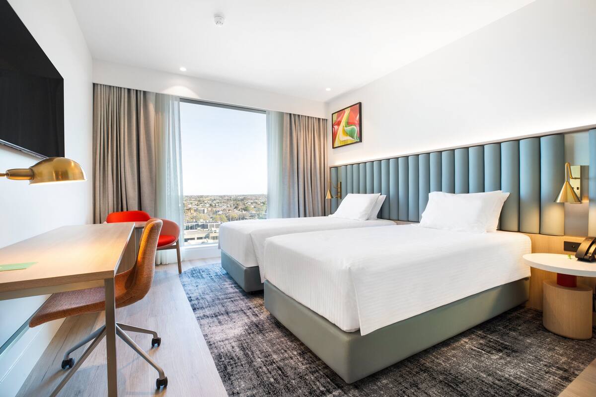 A hotel room with two beds, a desk, a chair, a small table, and a view of the city.