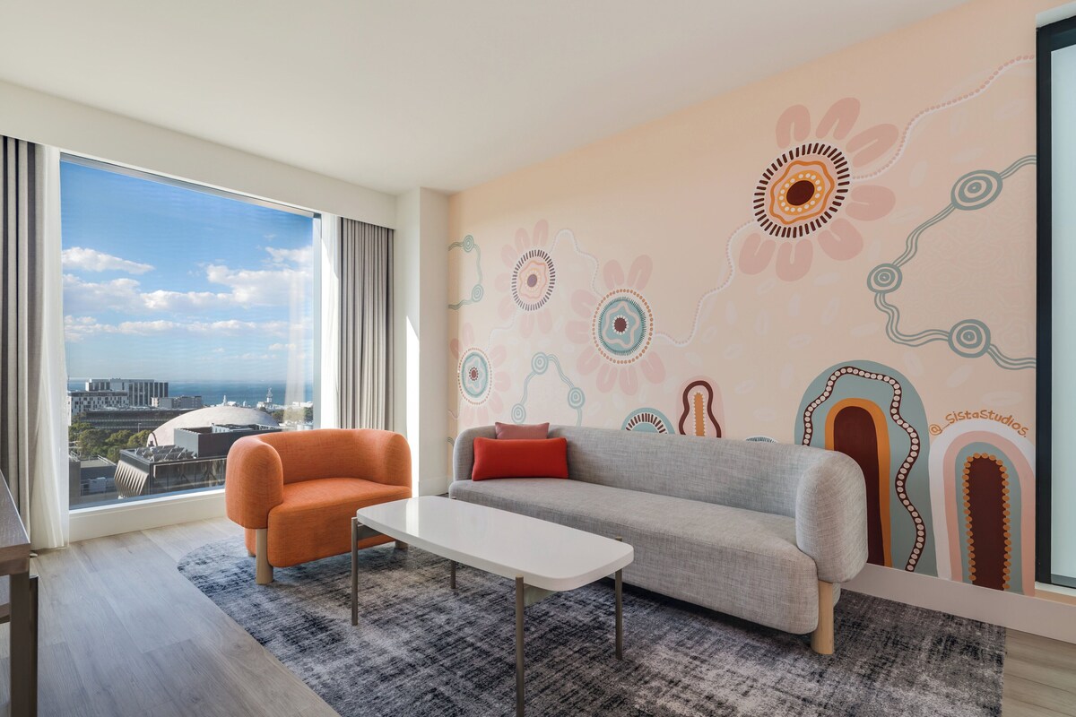 A modern living room with a sofa, armchair, and coffee table, large windows with a city view, and a decorative wall.