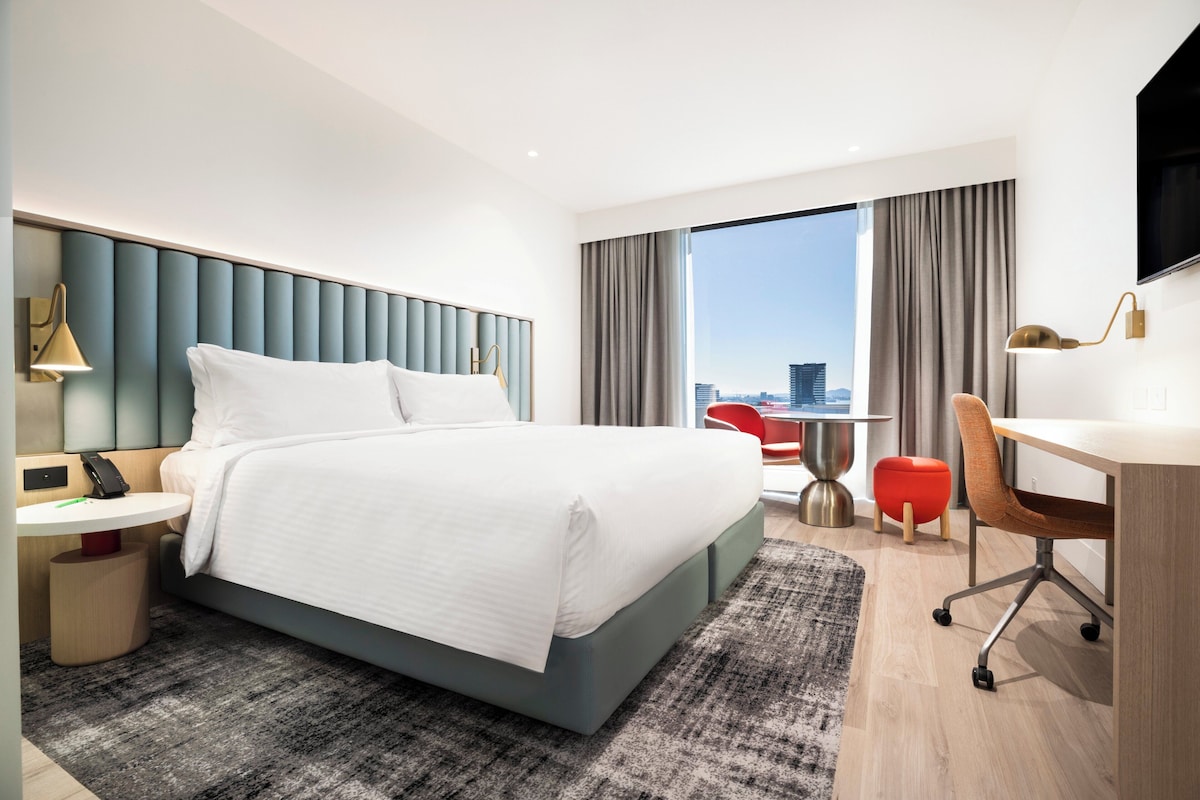 A modern hotel room with a large bed, a desk, a chair, and a view of the city.