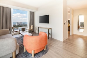 A modern hotel room with a large window, a flat-screen TV, a round table, and a seating area with a sofa and chairs.