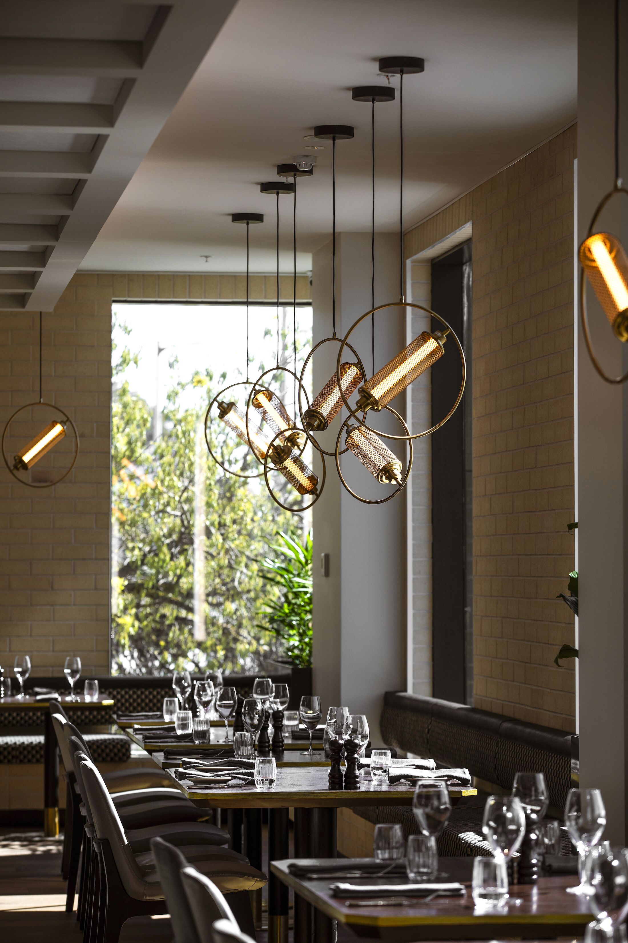 Un restaurante con varias mesas preparadas para comer, lámparas colgantes y vistas al exterior con vegetación.