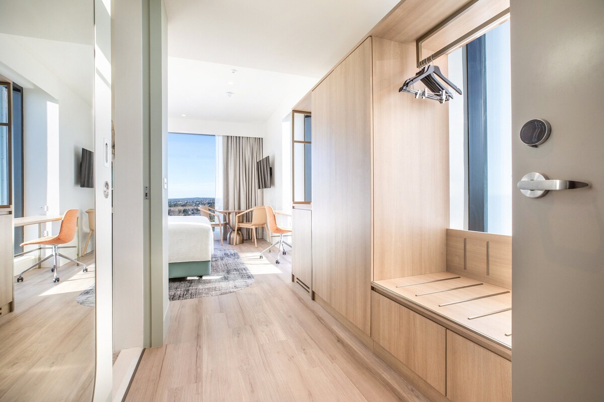 A modern hotel room with a wooden bench, a desk, and a large window offering a view.