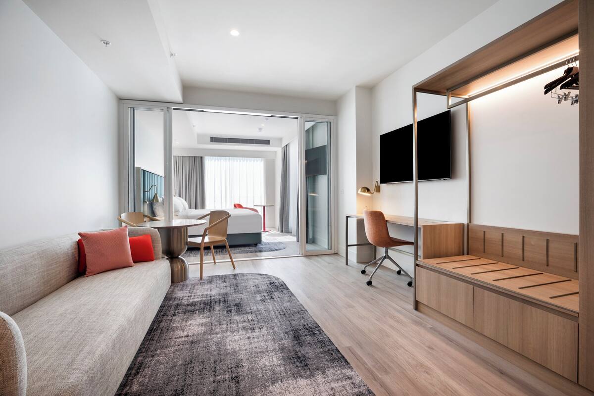 A modern hotel room with a sofa, a desk, a chair, a TV, and a wardrobe.
