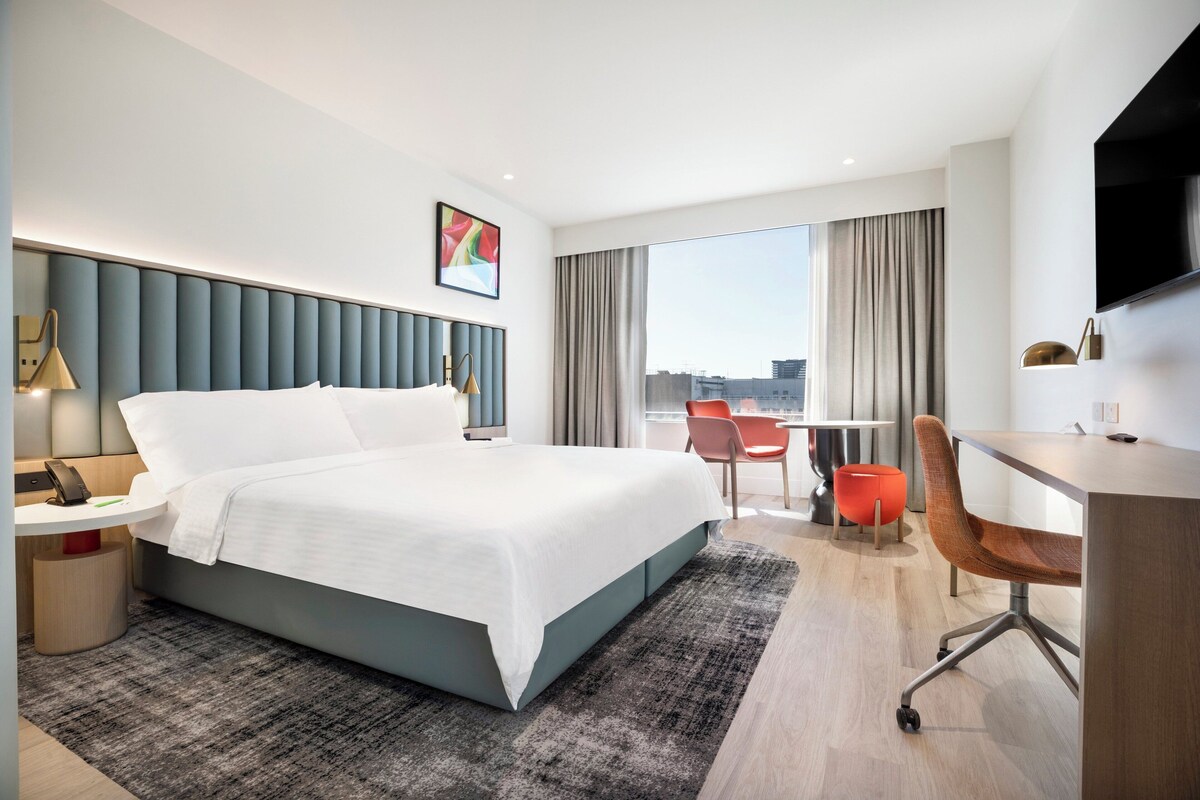A modern hotel room with a large bed, a desk, a chair, and a TV.