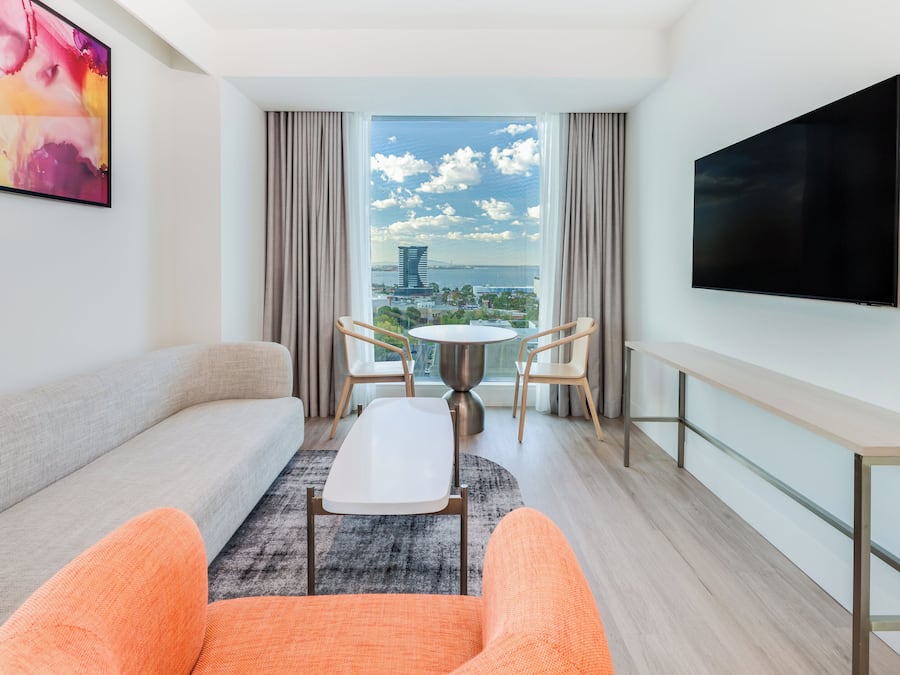 A modern hotel room with a sofa, two chairs, a small table, a TV, and a large window with a city view.