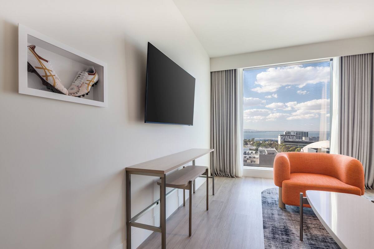 A modern hotel room with a large window, a flat-screen TV, a wooden desk, an orange armchair, and a framed pair of shoes on the wall.