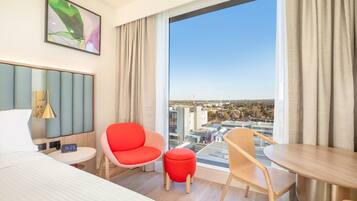 Habitación de hotel con cama, una mesa redonda, dos sillas, un pequeño reposapiés, una mesita de noche con lámpara, un reloj digital y un ventanal con vista a la ciudad.