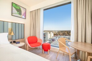 A hotel room with a bed, a round table, two chairs, a small ottoman, a bedside table with a lamp, a digital clock, and a large window with a city view. - Holiday Inn & Suites Geelong by IHG (Geelong)