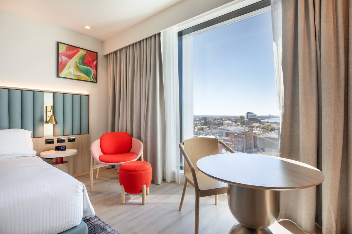 A modern hotel room with a large window, a bed, a red chair, a round table, and a city view.