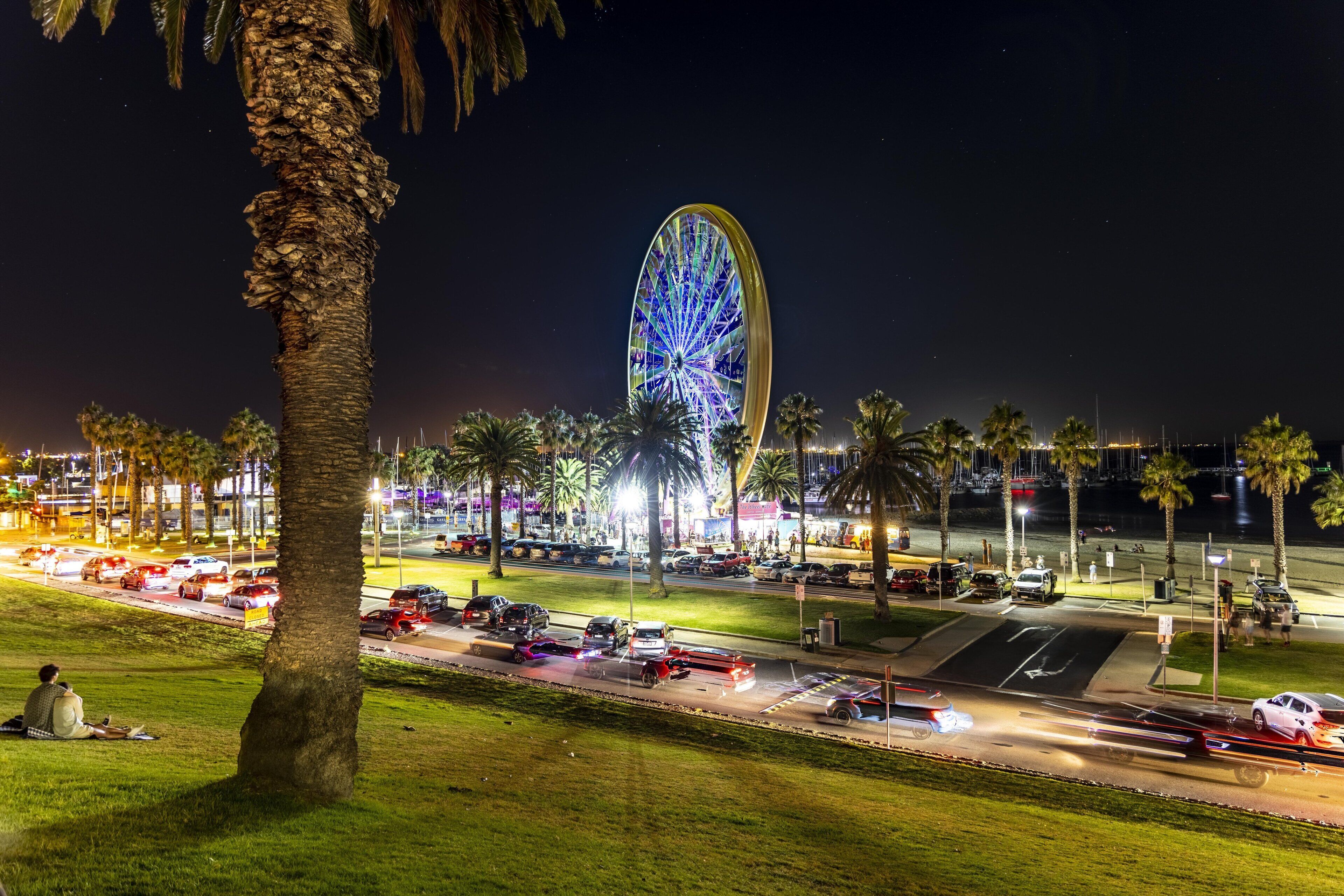 A night scene with a large illuminated Ferris wheel, palm trees, and a busy street with cars.