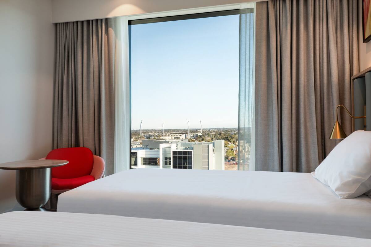 A hotel room with a large bed, a red chair, a small table, and a view of the city through a large window.