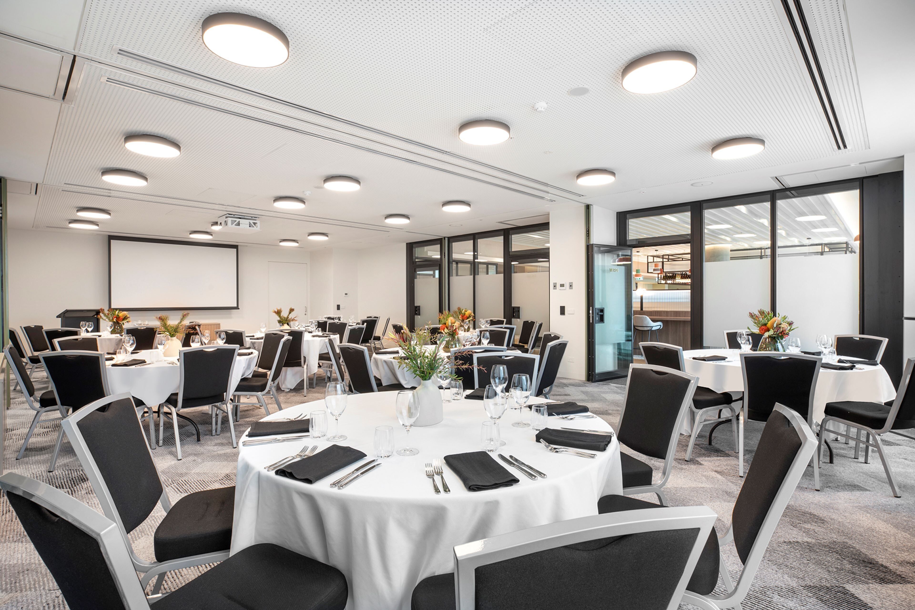 Een vergaderzaal met ronde tafels, zwarte stoelen en witte tafelkleden.