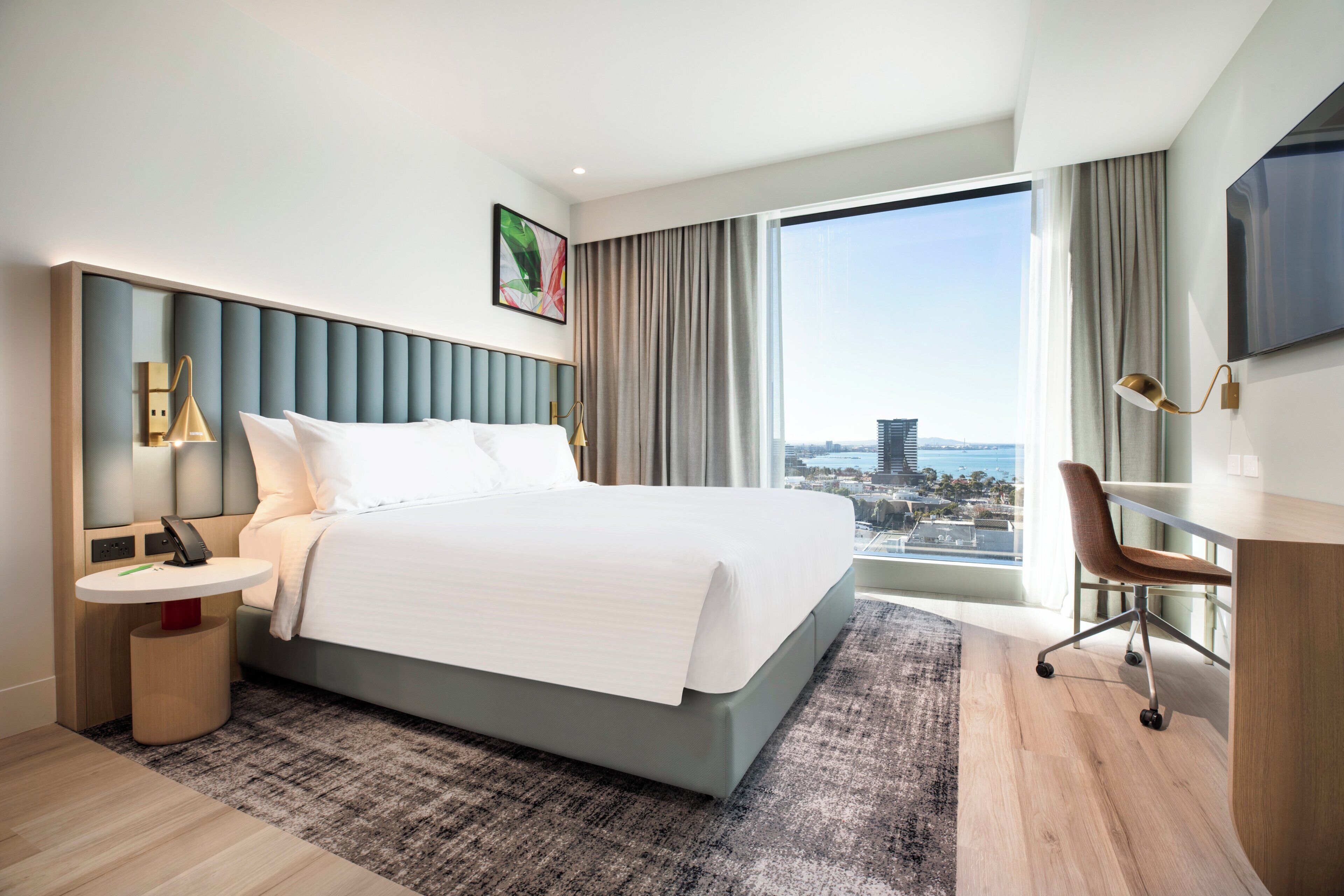 A hotel room with a large bed, a desk, a chair, a TV, and a view of the city.