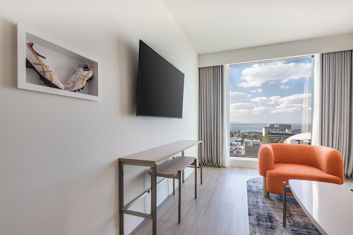 A modern hotel room with a large window, a flat-screen TV, a wooden desk, an orange armchair, and a framed pair of shoes on the wall.