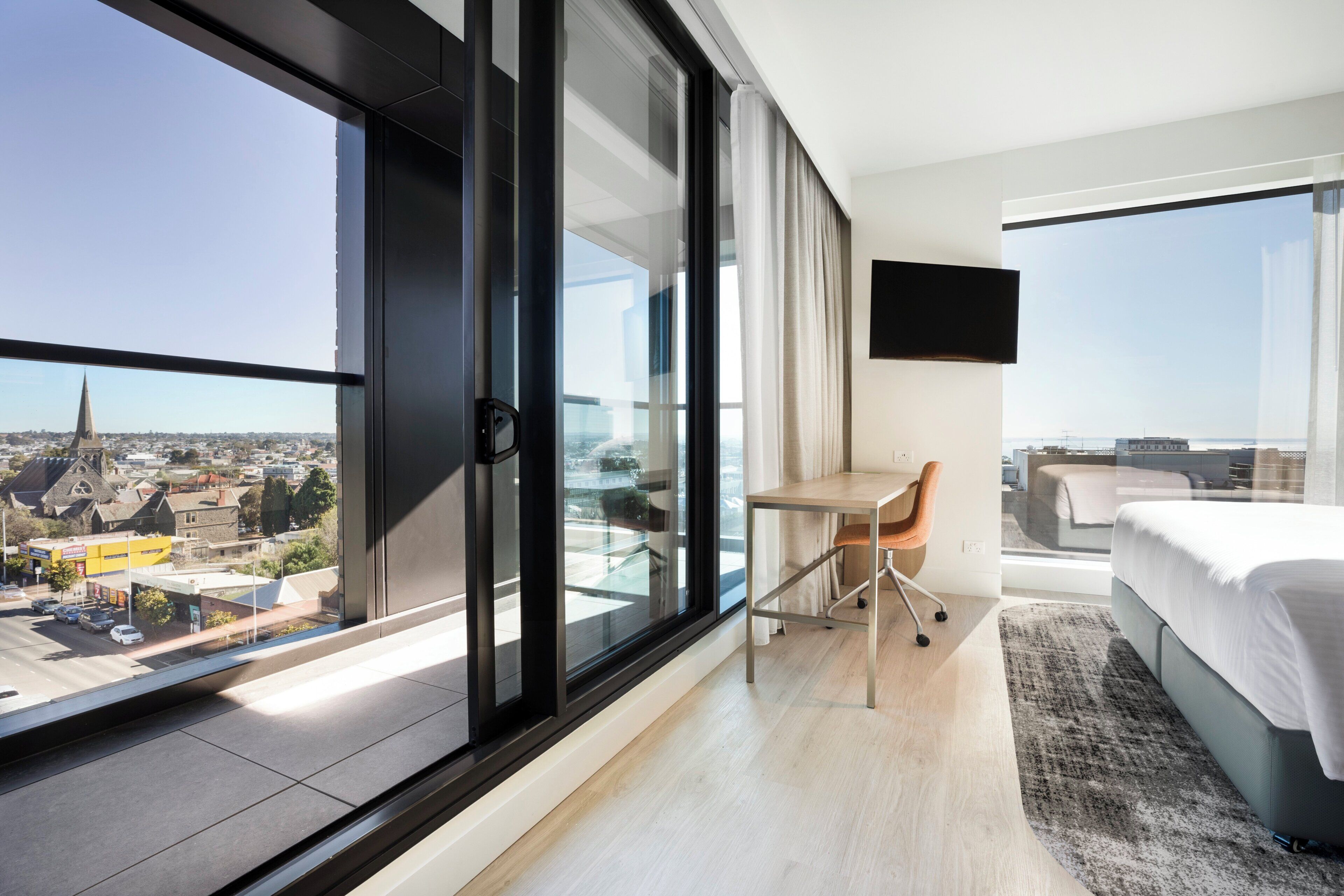 Una habitación de hotel moderna con un amplio ventanal corredero, una cama, un escritorio y un televisor.