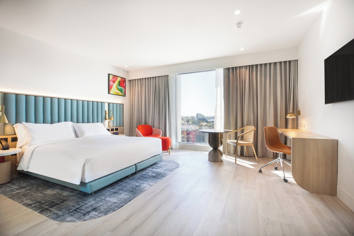 A modern hotel room with a large bed, a desk, and a view of the city.