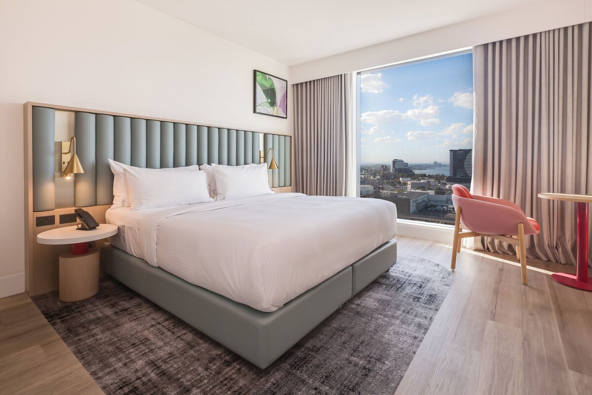 A modern hotel room with a large bed, a city view, a red chair, and a small table.