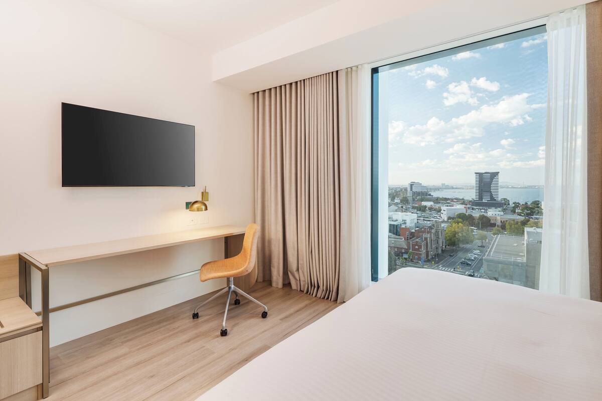 A modern hotel room with a large bed, a desk with a chair, a flat-screen TV, and a large window with a city view.