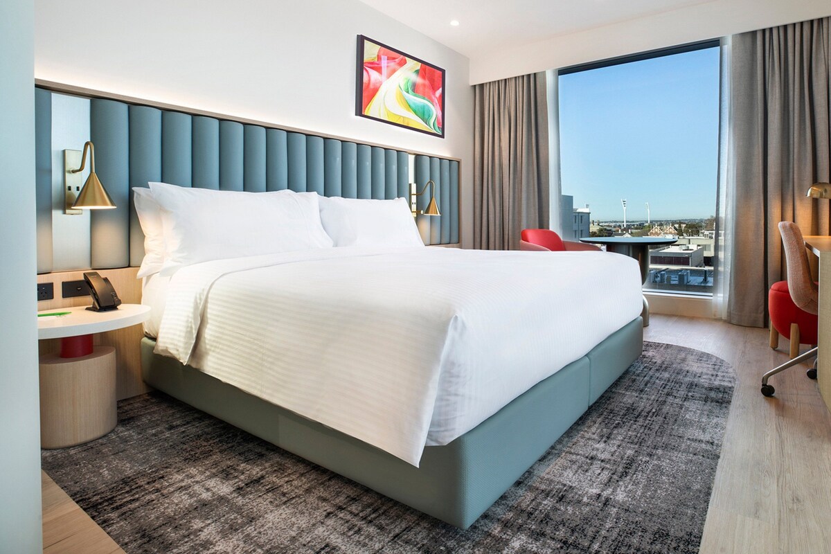 A modern hotel room with a large bed, a desk, a chair, and a view of the city.