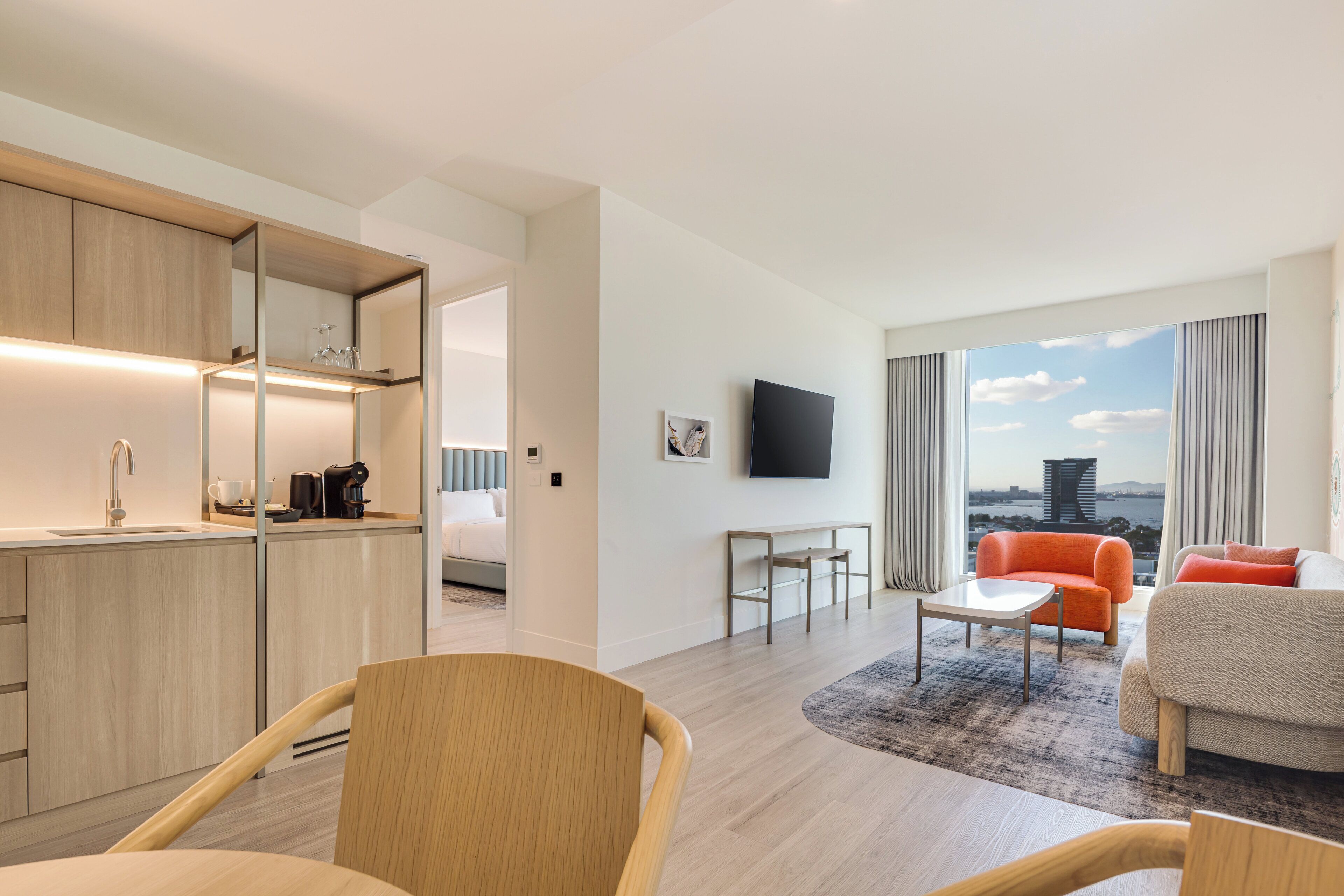 Quarto de hotel moderno com cozinha, área de refeições, sala de estar com sofá e poltrona, e uma janela grande com vista para a cidade.