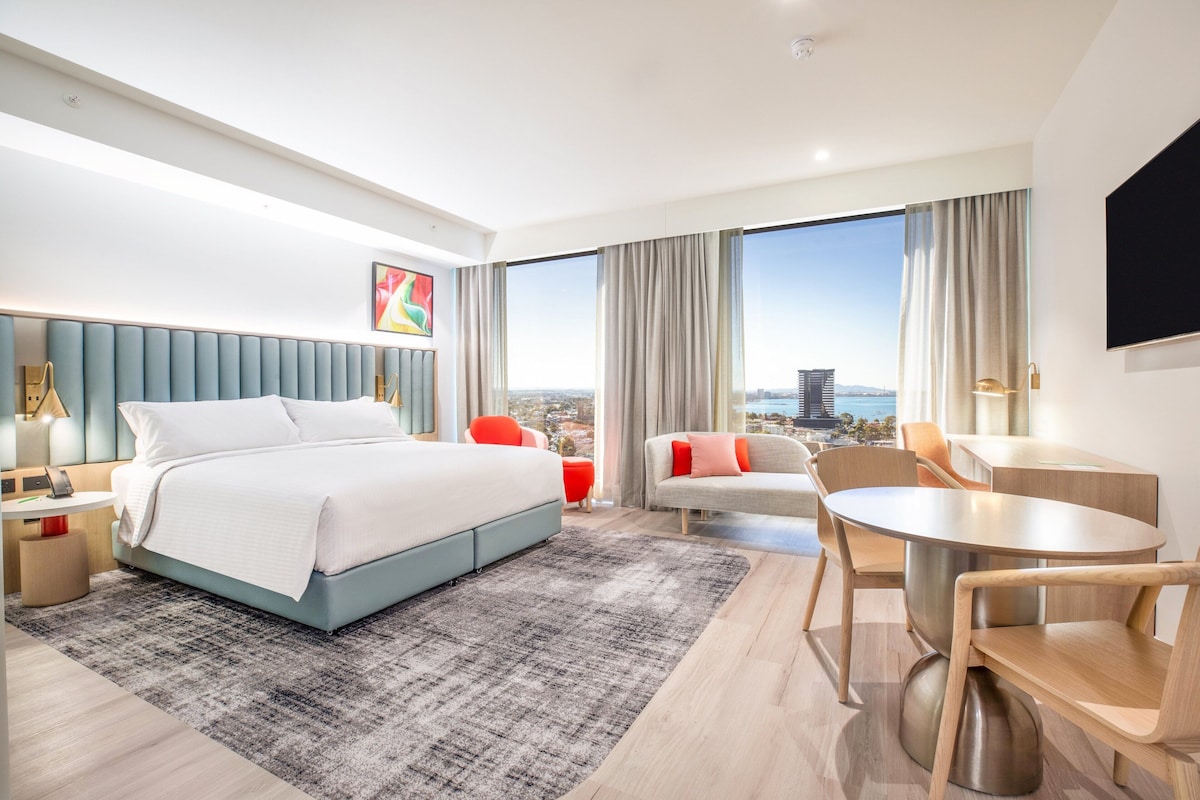 A modern hotel room with a large bed, a seating area with a table, and a view of the cityscape through large windows.